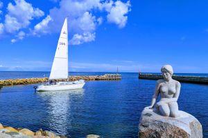 Eine Statue einer Meerjungfrau, die auf einem Felsen neben einem Segelboot sitzt.