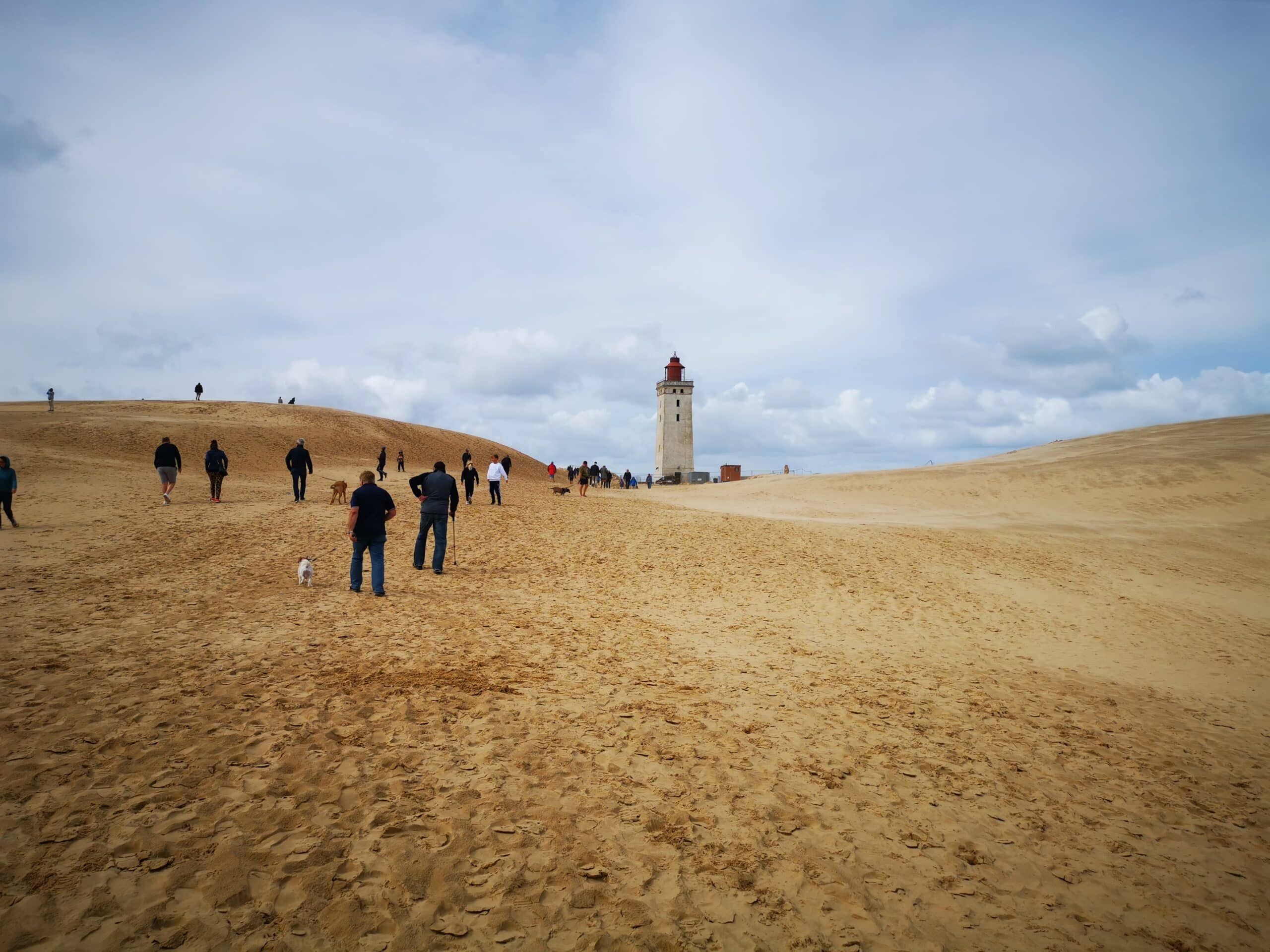 Eine Gruppe von Menschen, die auf einer Sanddüne in der Nähe eines Leuchtturms spazieren gehen.