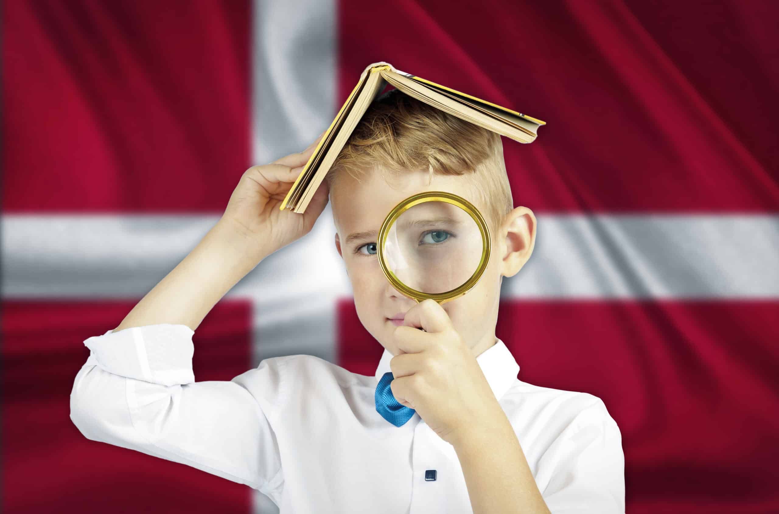 kleiner Junge mit Lupe in der Hand und Buch auf dem Kopf vor Dänemarks Fahne