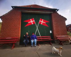Zwei Menschen und ein Hund vor einem roten Gebäude mit aufgemalten dänischen Flaggen.