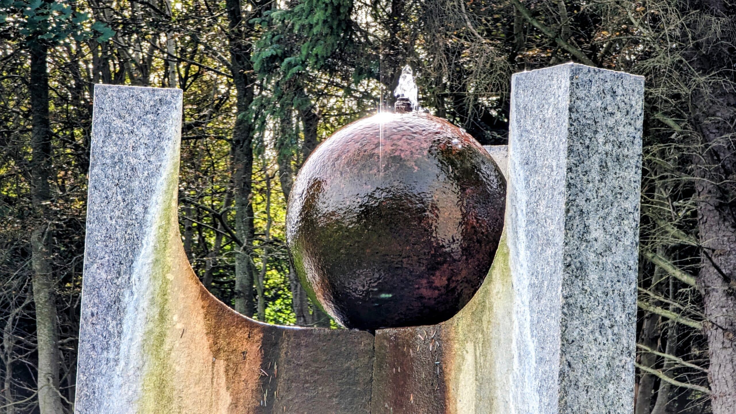 Eine Skulptur mit einer Metallkugel mitten in einem Waldgebiet.